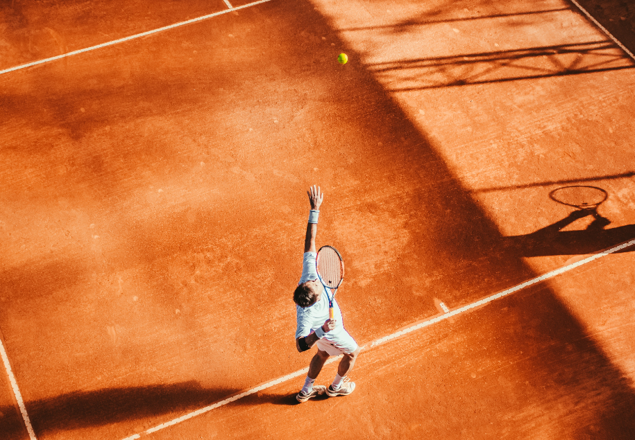 Tenisista na korcie tenisowym, widoczny z góry, podrzuca piłeczkę nad głowę aby zaserwować